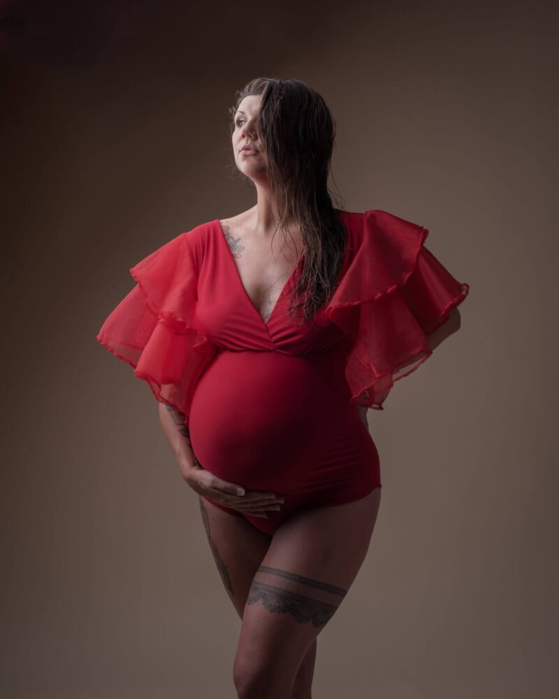 Pregnant women posed in red maternity bodysuit holding bump